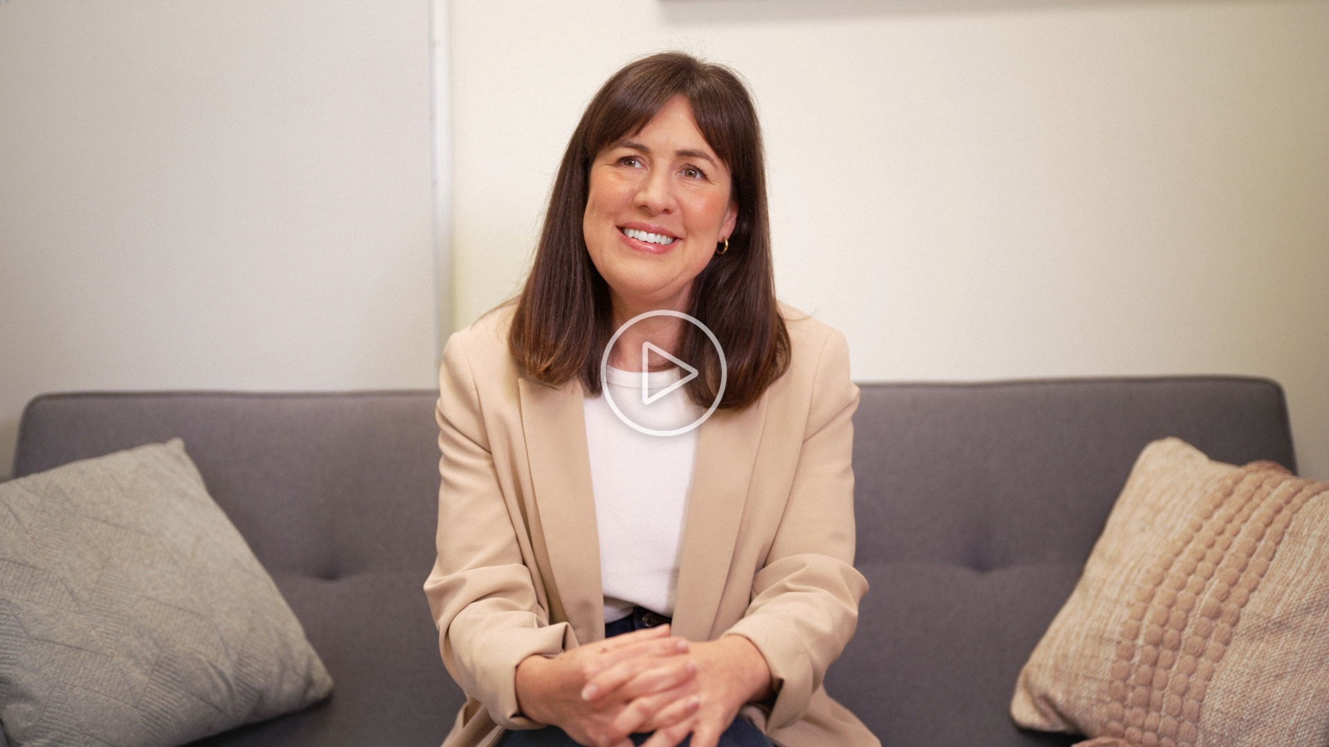 Associate Professor Lou Farrer smiling while seated on a grey couch, wearing a beige blazer and white top, with cushions on either side. 